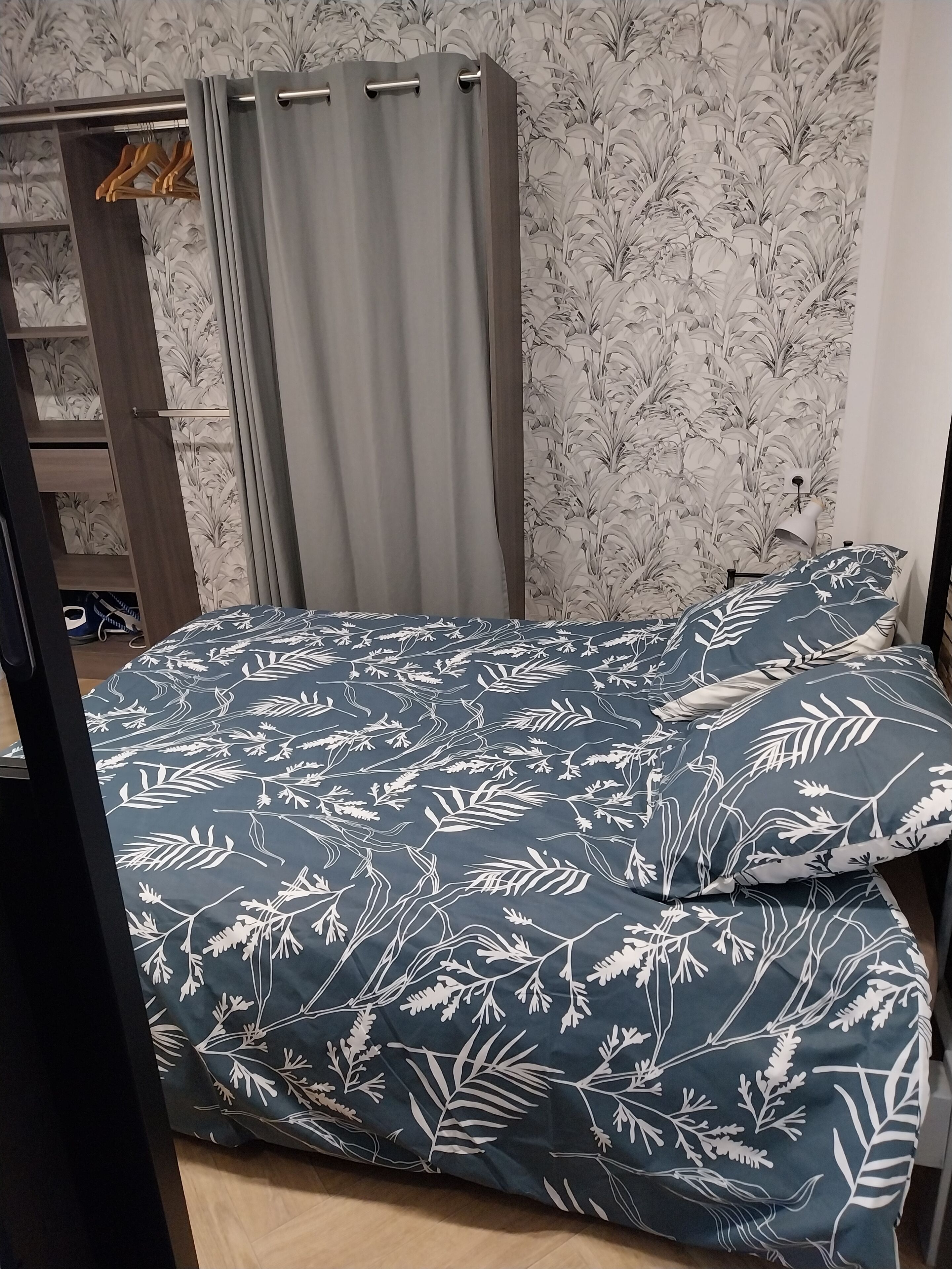 Une chambre à coucher avec un lit recouvert d’une couvre-lit à motifs floraux bleu foncé, assorti aux coussins, une armoire en bois et un rideau gris.