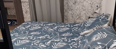 Une chambre à coucher avec un lit recouvert d’une couvre-lit à motifs floraux bleu foncé, assorti aux coussins, une armoire en bois et un rideau gris.
