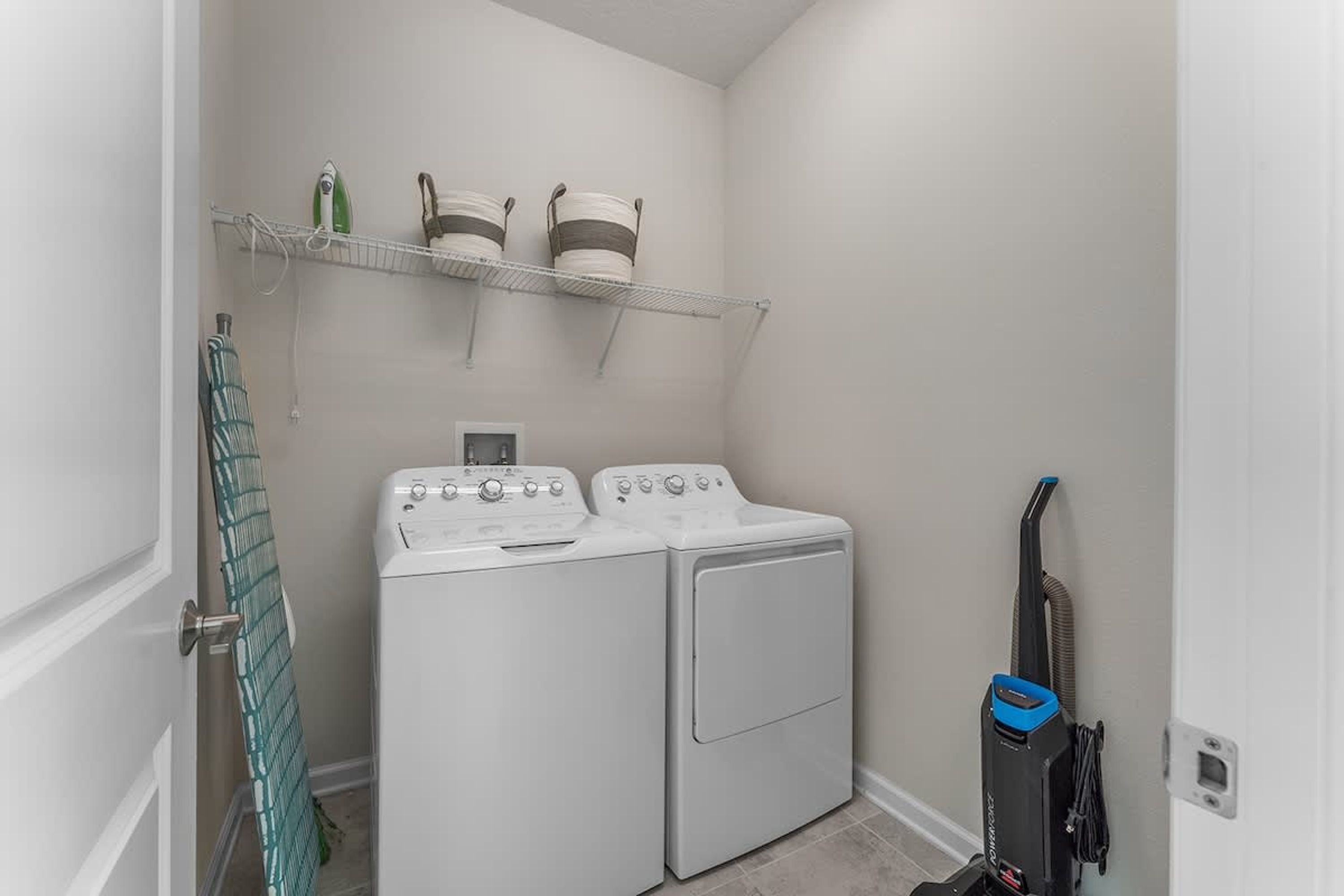 A laundry room with a washer and dryer, a shelf with laundry supplies, and a vacuum cleaner.