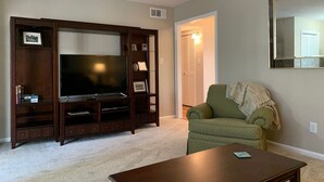 Una sala de estar con un televisor de pantalla plana grande sobre un mueble de madera, un sillón verde y una mesa de centro.