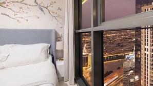 Un dormitorio con una cama, una mesita de noche, una lámpara y una ventana con vistas a la ciudad.