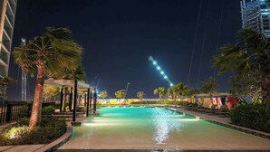 Un área de piscina nocturna con palmeras, sillas de descanso y un paseo bien iluminado.