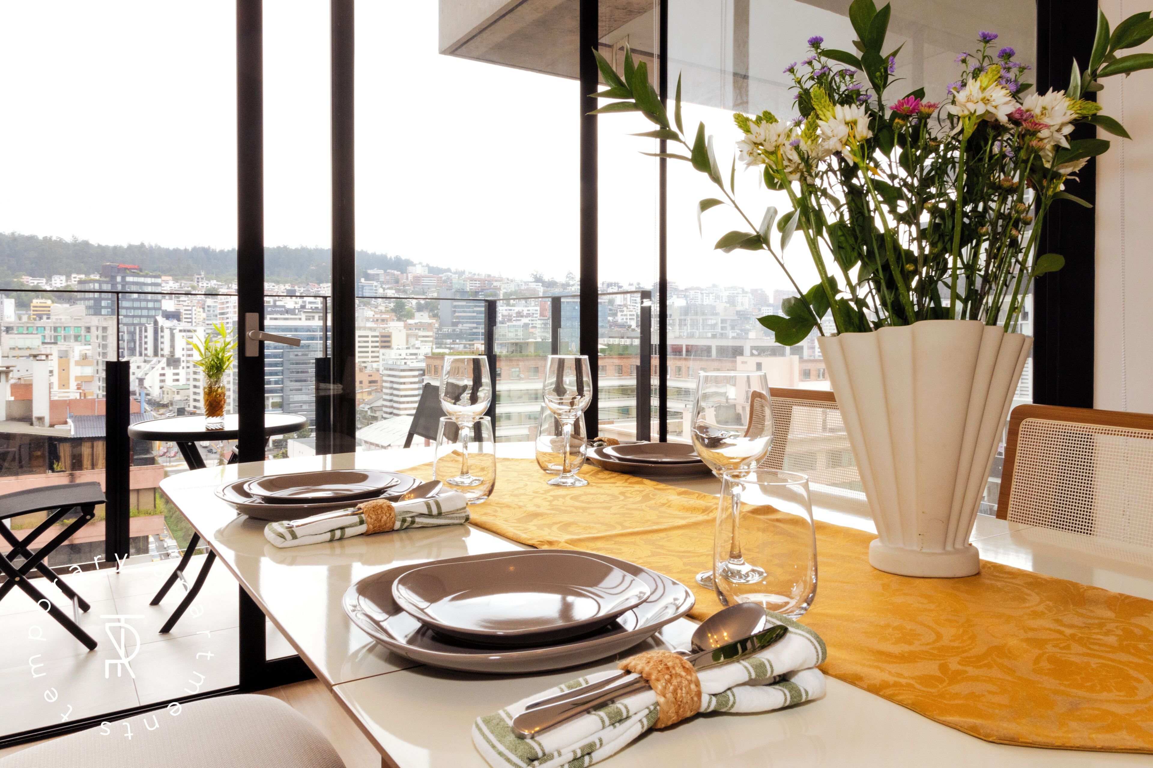 A dining table set for four with plates, cutlery, and glasses, overlooking a cityscape through large windows.