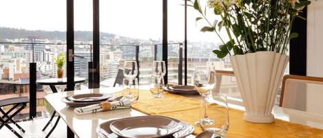 A dining table set for four with plates, cutlery, and glasses, overlooking a cityscape through large windows.