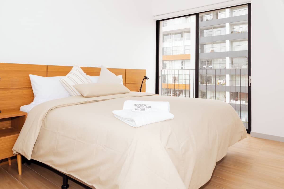 A neatly made bed with a beige comforter and pillows, a wooden headboard, a bedside table, and a view of a modern building through a large window.