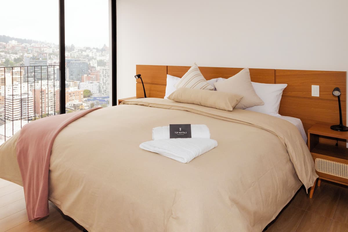 A neatly made bed with a wooden headboard, a bedside table with a lamp, and a view of a cityscape through large windows.