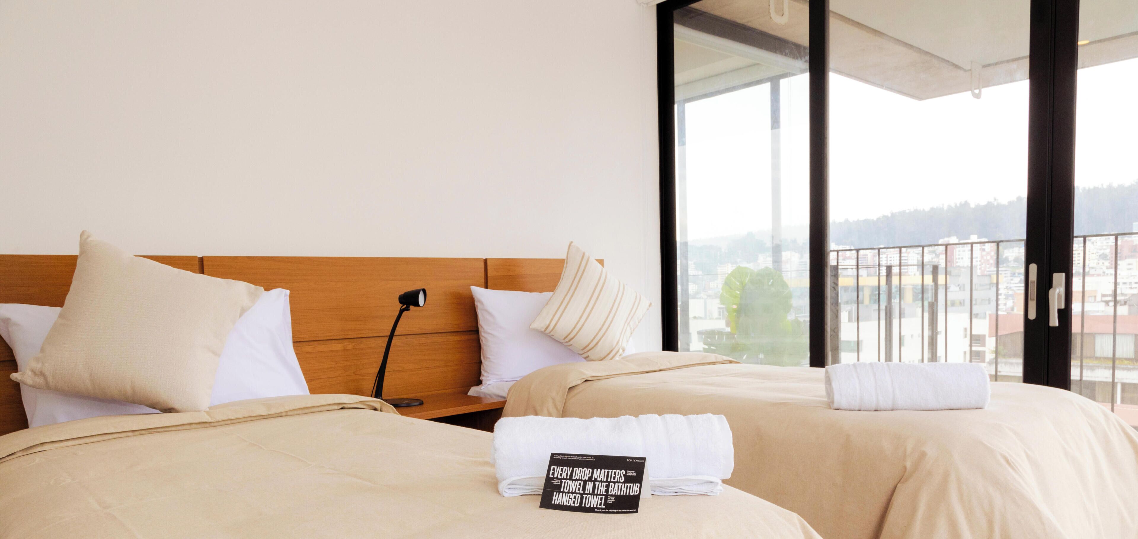 Ein Hotelzimmer mit zwei Betten, einem Nachttisch mit Lampe und Blick auf die Stadt durch große Fenster.