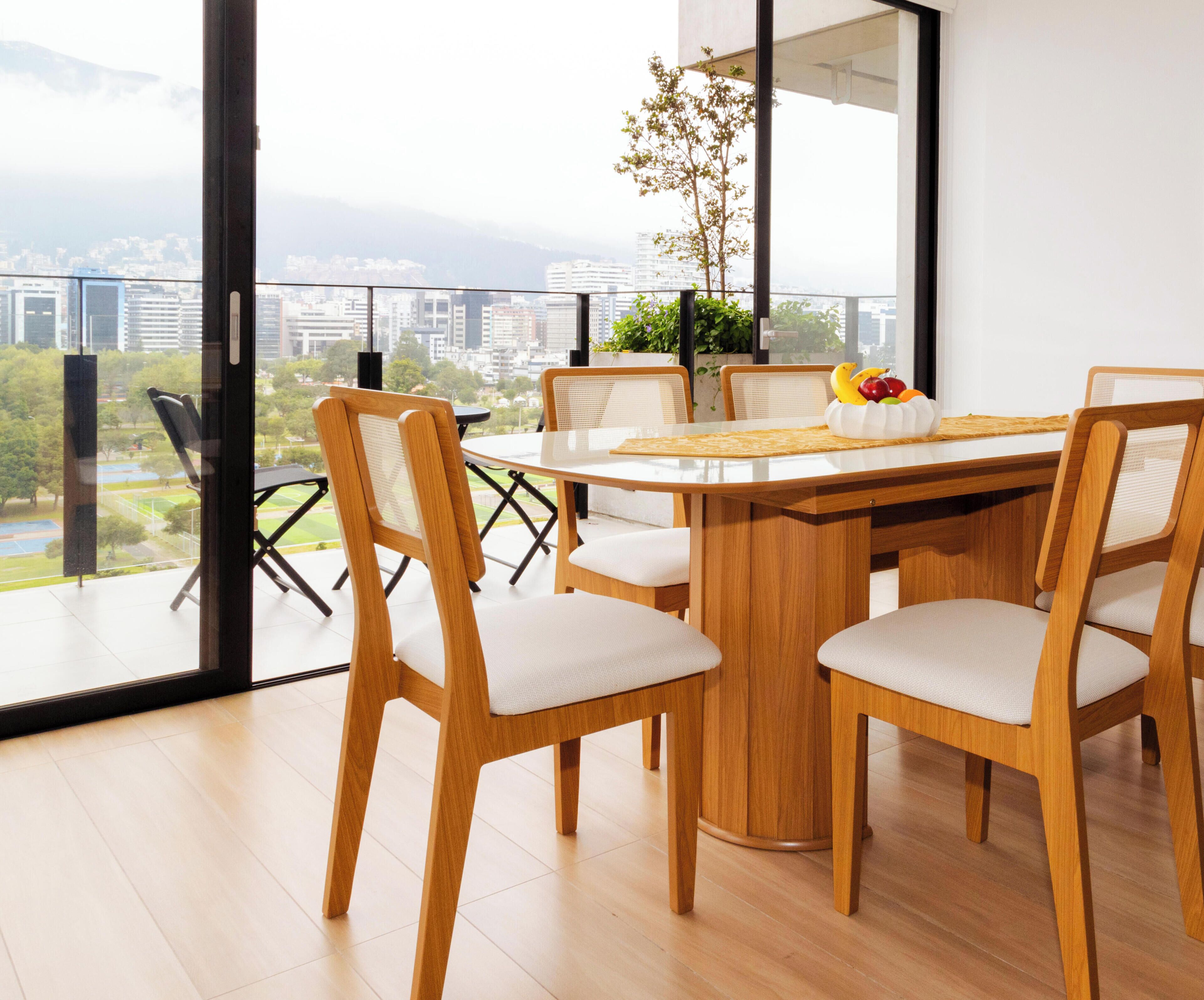 Un comedor moderno con una mesa redonda, seis sillas de madera con asientos acolchados y vista a un paisaje urbano a través de amplias ventanas.
