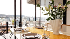 Una mesa de comedor lista con platos, vasos y un jarrón de flores, con vista a un paisaje urbano.