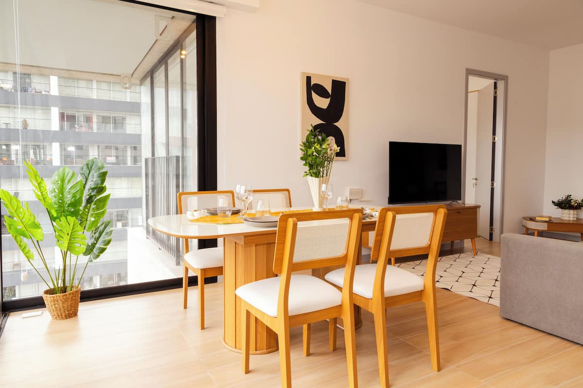 A modern living room with a dining area, a flat-screen TV, and a large window with a view of a building.