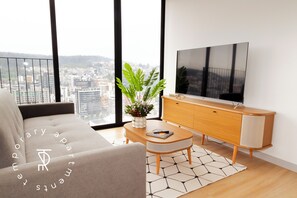 Un salon moderne avec un canapé, un téléviseur posé sur un meuble en bois, une plante et une vue sur la ville à travers de grandes fenêtres.