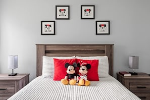Quarto com cama, dois travesseiros do Mickey Mouse, cabeceira, duas mesas de cabeceira e quadros nas paredes.