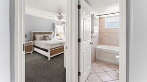 A bedroom with a bed, nightstands, a ceiling fan, and a view of a bathroom with a bathtub and toilet.