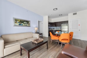 A living room with a beige sofa, a coffee table, and orange chairs. There is a kitchen in the background with a microwave, stove, and cabinets. - Hotel Apt at Celebration 10 min From Disney (Celebration)