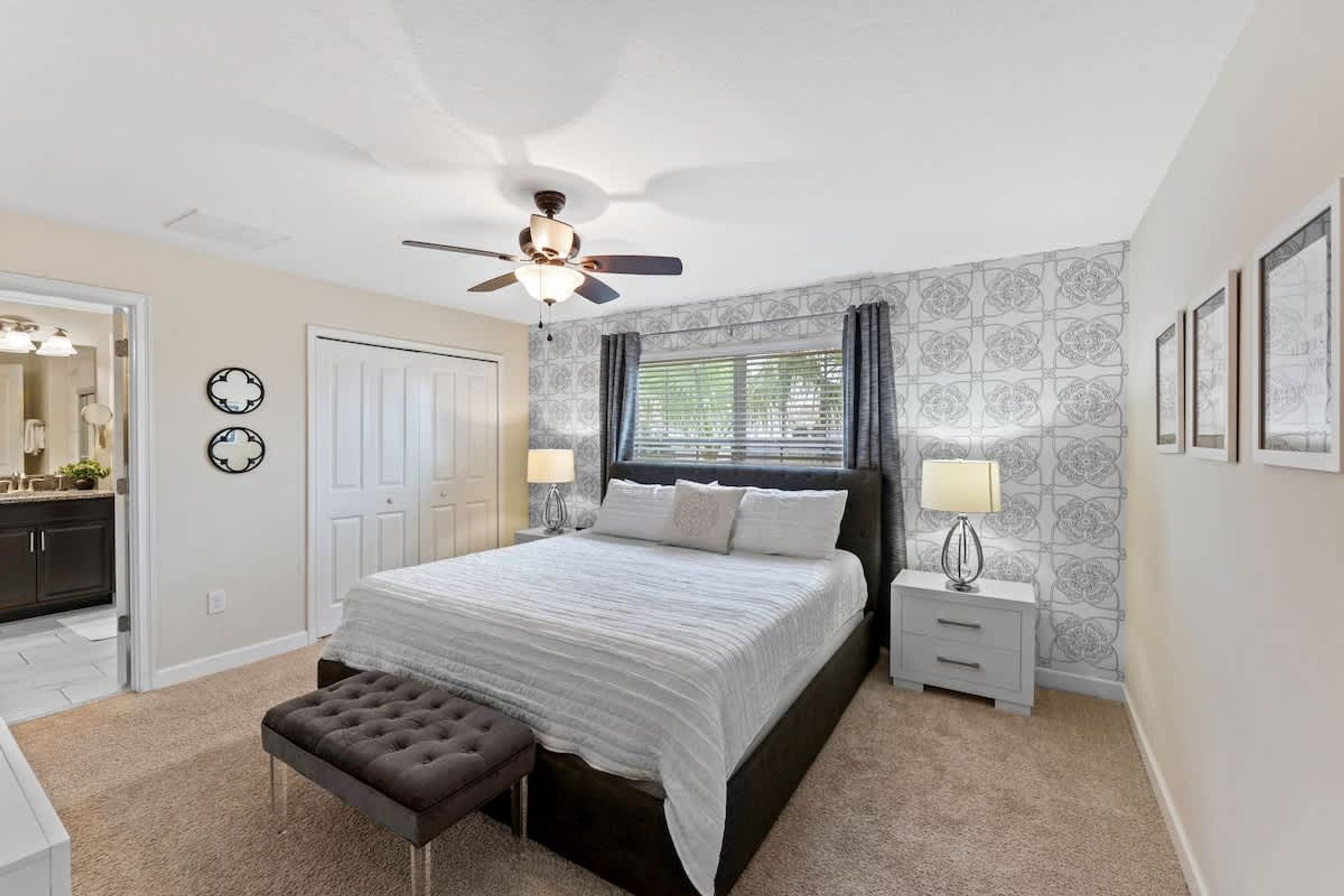 A bedroom with a bed, a ceiling fan, a nightstand, a lamp, a bench, and a door leading to another room.