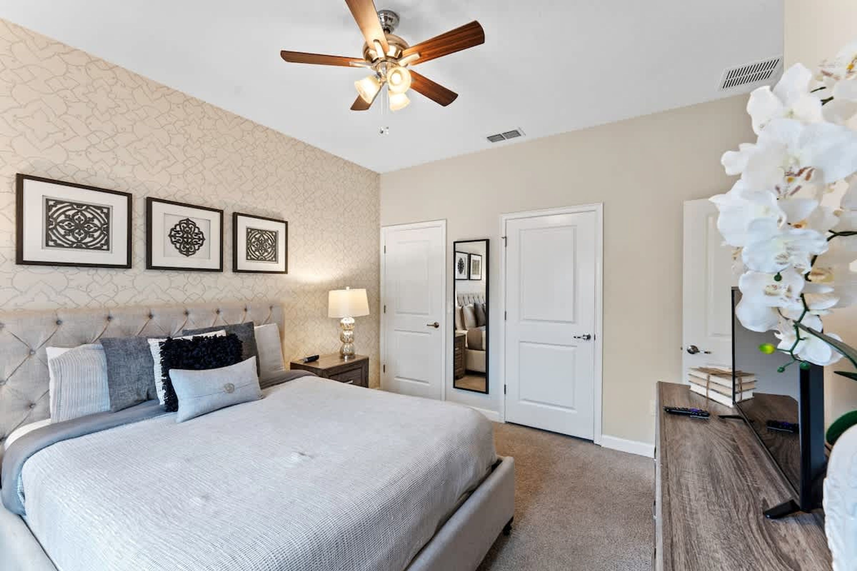 A bedroom with a bed, a ceiling fan, framed artwork on the wall, a mirror, and a white orchid plant.