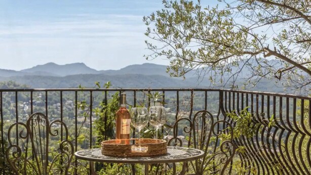 A balcony with a table set for two, featuring a bottle and glasses, overlooking a mountainous landscape.