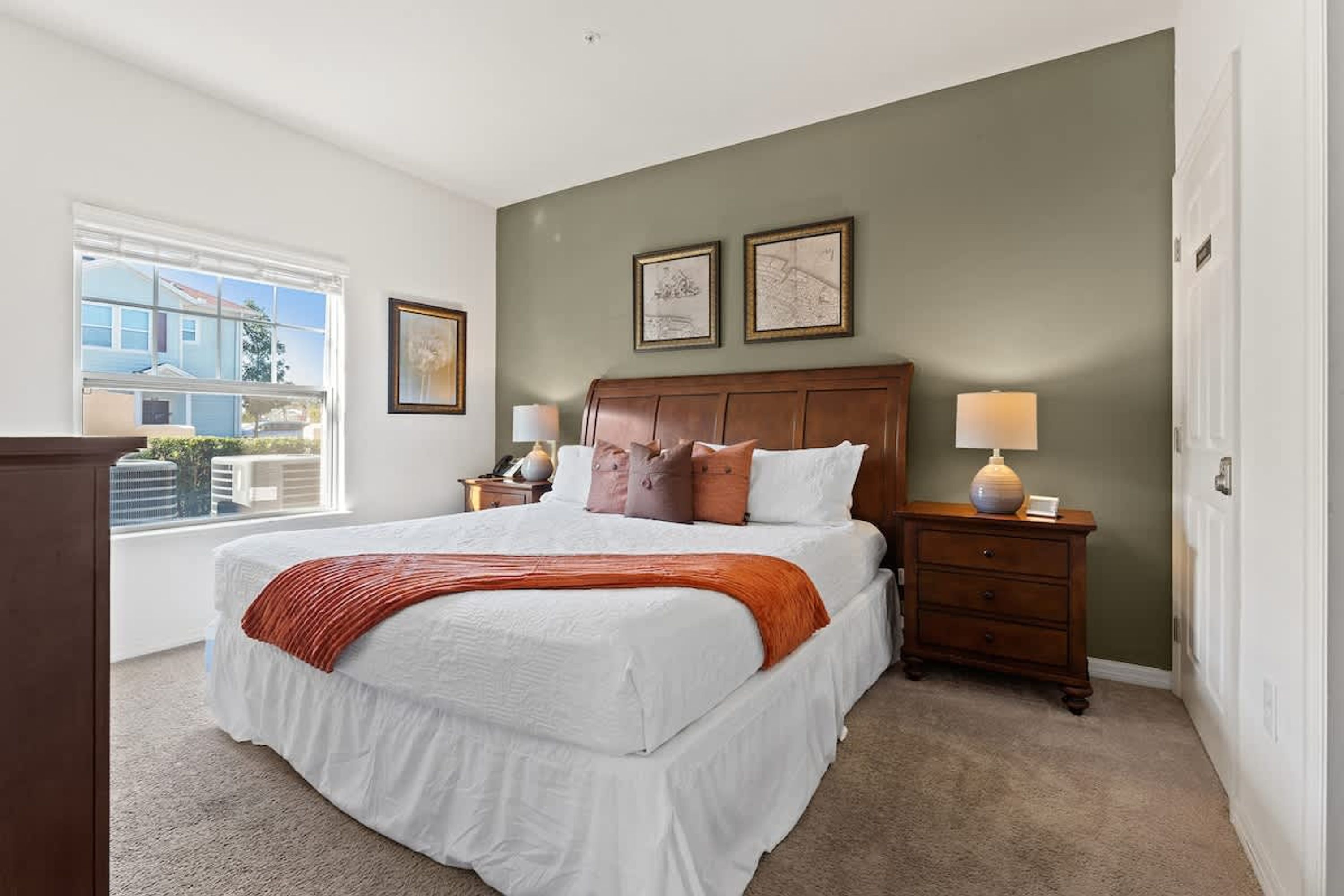 A bedroom with a large bed, two bedside tables with lamps, a window with a view, and two framed pictures on the wall.