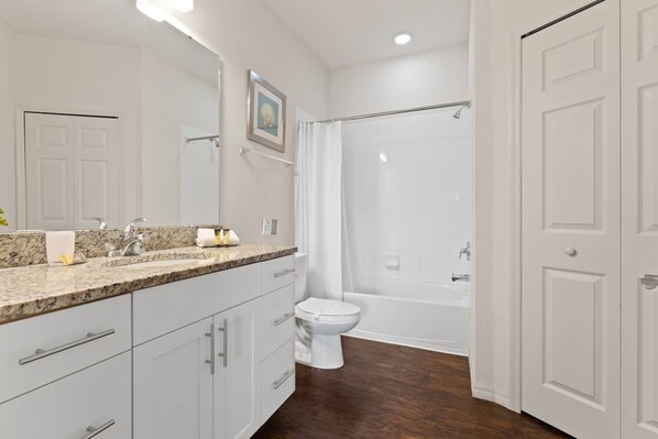 Une salle de bain moderne avec un lavabo surmonté d’un plan de travail en granit, des toilettes blanches, une baignoire et un rideau de douche.