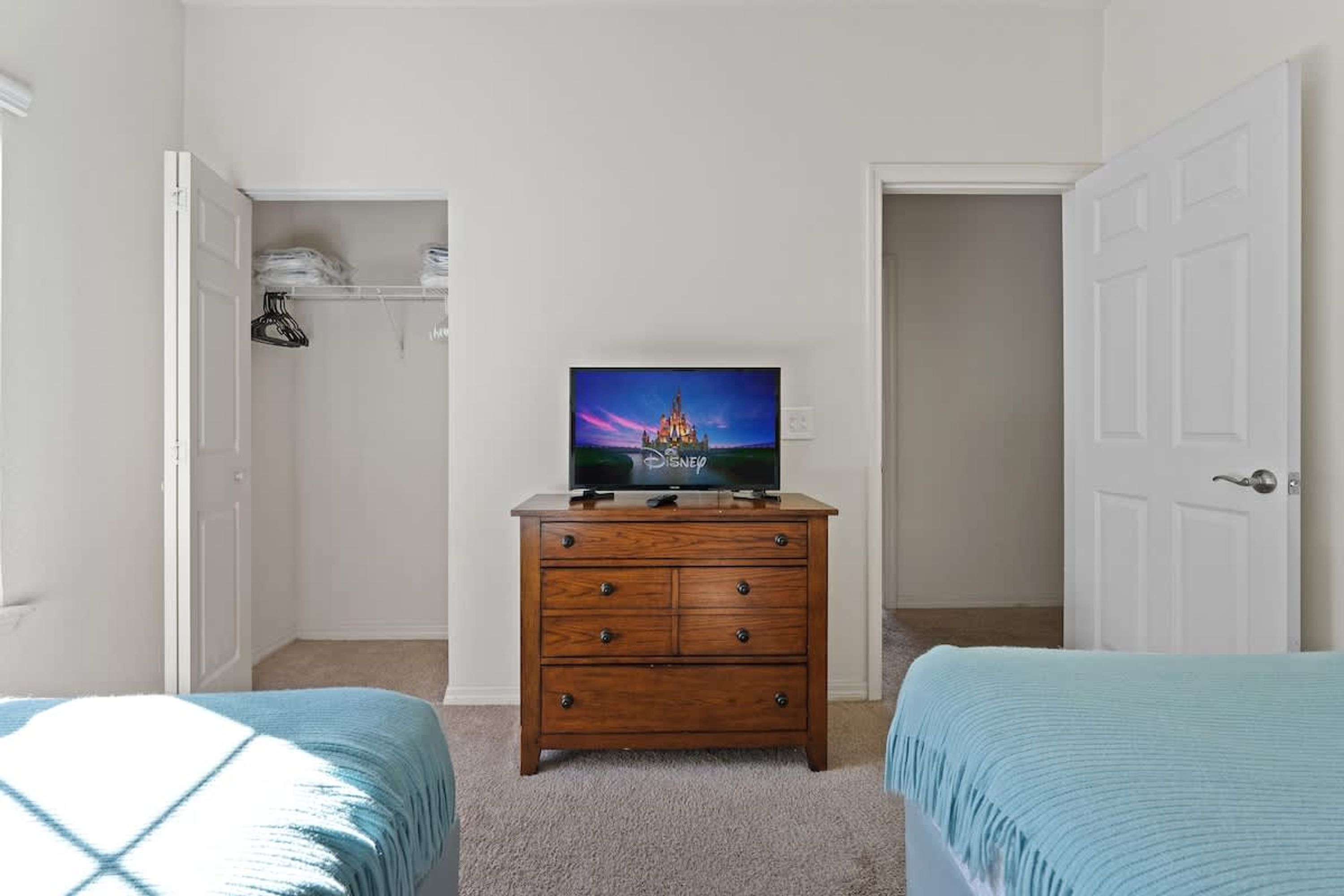 A bedroom with two beds, a wooden dresser, a television on the dresser, and a closet with hanging clothes.