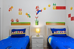 A bedroom with two beds, each with a blue bedspread featuring Mario characters. There are wall decals of Mario, Luigi, and other characters. A nightstand with a lamp and a small Mario figurine is between the beds. - Game Room Plus Private Pool 4BR Disney Area (Kissimmee)