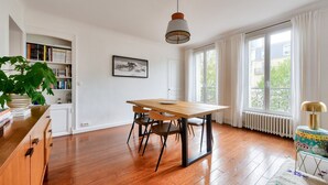 Une pièce spacieuse avec un plancher en bois, une table à manger en bois avec des chaises, une bibliothèque, un tableau encadré au mur et de grandes fenêtres avec des rideaux.