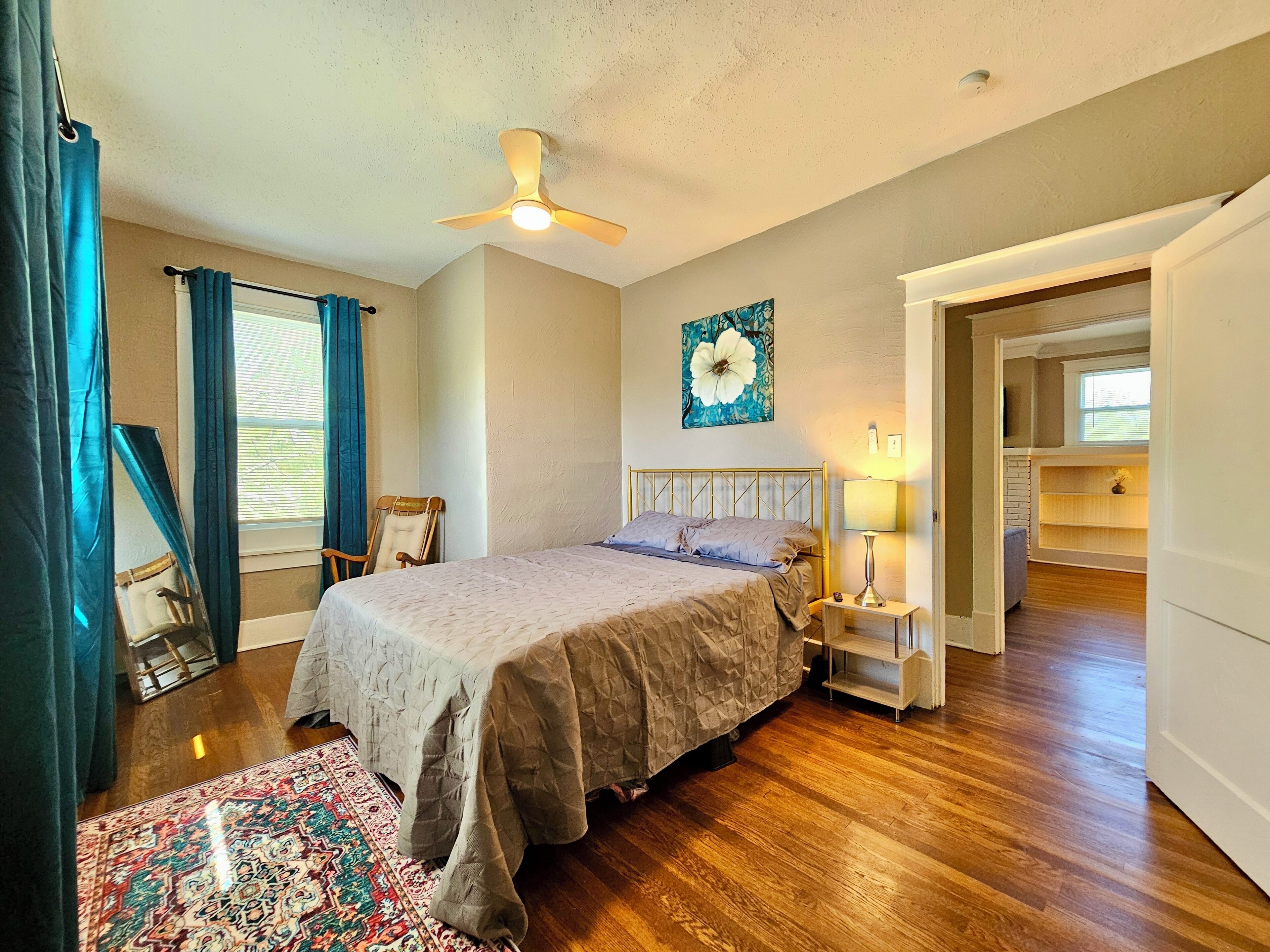 A bedroom with a bed, a ceiling fan, a nightstand with a lamp, a large floral painting on the wall, and a view into an adjacent room.