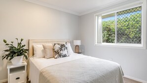 Ein Schlafzimmer mit Bett, Nachttisch, Zimmerpflanze und Fenster mit Blick auf Grünflächen.