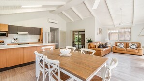 Eine geräumige Küche mit einer großen Kochinsel, ein Essbereich mit Holztisch und weißen Stühlen sowie ein Wohnzimmer mit braunem Ledersofa und einem großen Fenster.