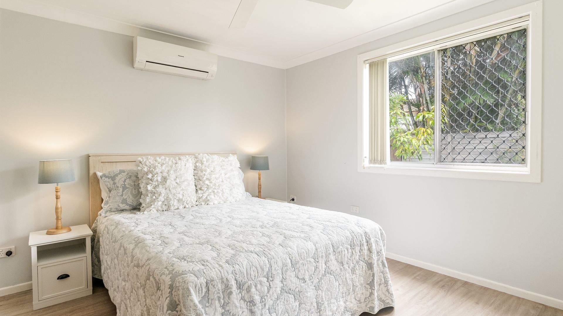 Quarto com cama, mesas de cabeceira, luminárias, ar condicionado e janela com vista para o verde.
