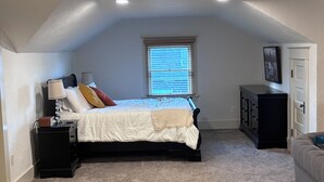 A bedroom with a bed, bedside tables, a window with blinds, a dresser, and a door. - UPS Neighborhood Apartment (Tacoma)