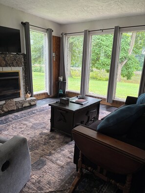 A living room with a fireplace, large windows, a patterned rug, and a wooden bench. - Charming 2-bedroom bungalow with AC, WiFi,washer and dryer, ON THE CREEK. (Ellwood City)
