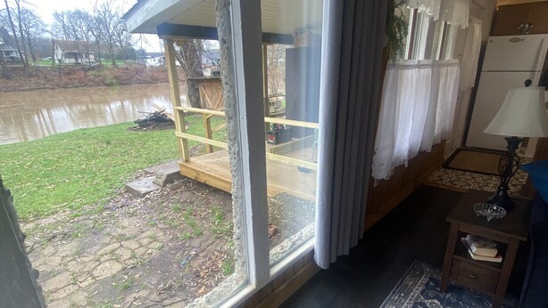 A room with a large window overlooking a yard and a body of water. There is a wooden deck outside the window, a rug on the floor, and a lamp on a side table.