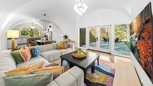 Un vaste salon comprenant un grand canapé, une table basse et un tableau représentant une cascade. On aperçoit une cuisine à travers une arche, ainsi qu’une table à manger avec des chaises.