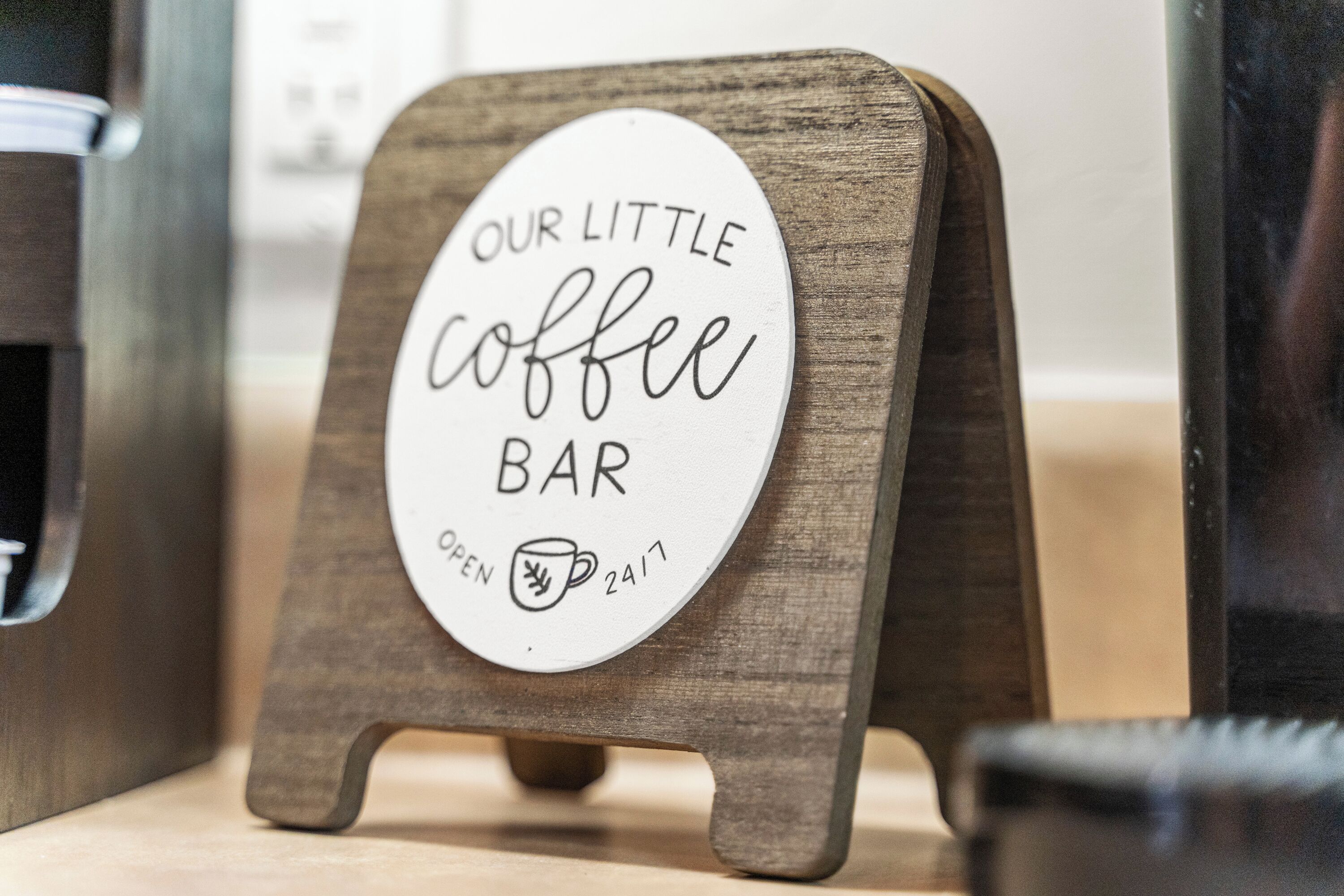 A sign on a wooden stand that reads "Our Little Coffee Bar" with an open icon and a coffee cup, indicating the bar is open 24 hours.