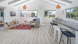 A spacious living area with a high ceiling, large windows, and a kitchen area with bar stools.