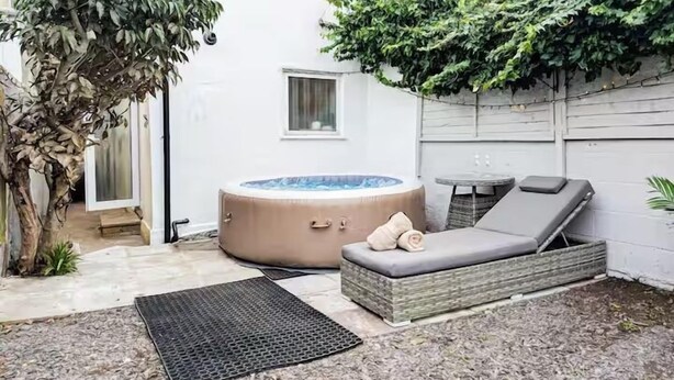 A hot tub with a lounge chair and cushions, surrounded by a small garden and a white wall.