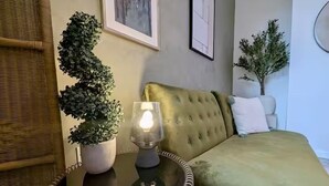 A room with a green upholstered bed, a round side table with a lamp, and two potted plants.