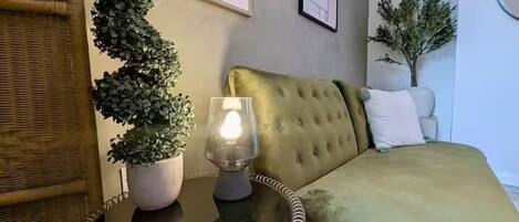 Una habitación con una cama tapizada de verde, una mesita redonda con una lámpara y dos plantas en macetas.