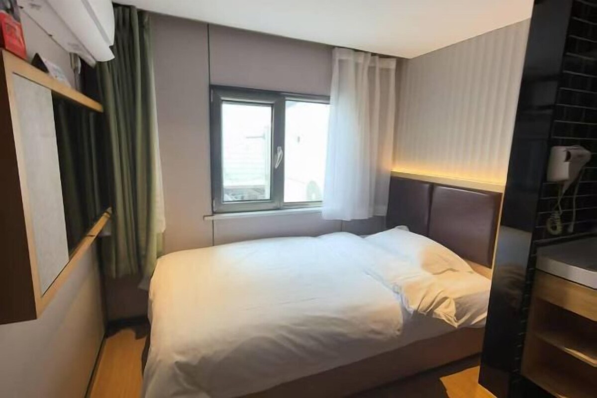 A single bed with a headboard, a window with curtains, a built-in shelf, and a wall-mounted air conditioning unit.