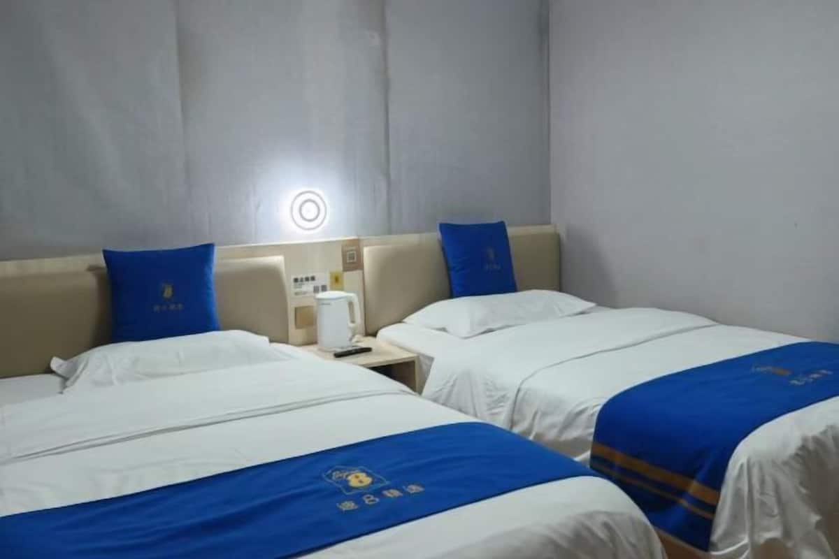 A hotel room with two beds, each with a blue bedspread and matching pillows. There is a nightstand with a white lamp and a small table with a white kettle.