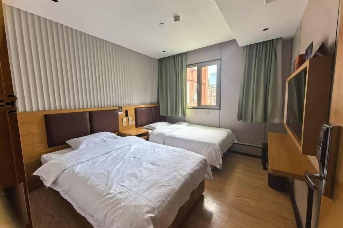 A hotel room with two beds, a wooden headboard, a small table, a TV, and a window with curtains.