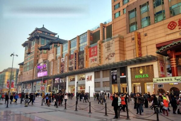 A busy urban shopping area with modern and traditional architecture, featuring prominent retail stores like Zara, H&M, and Rolex.