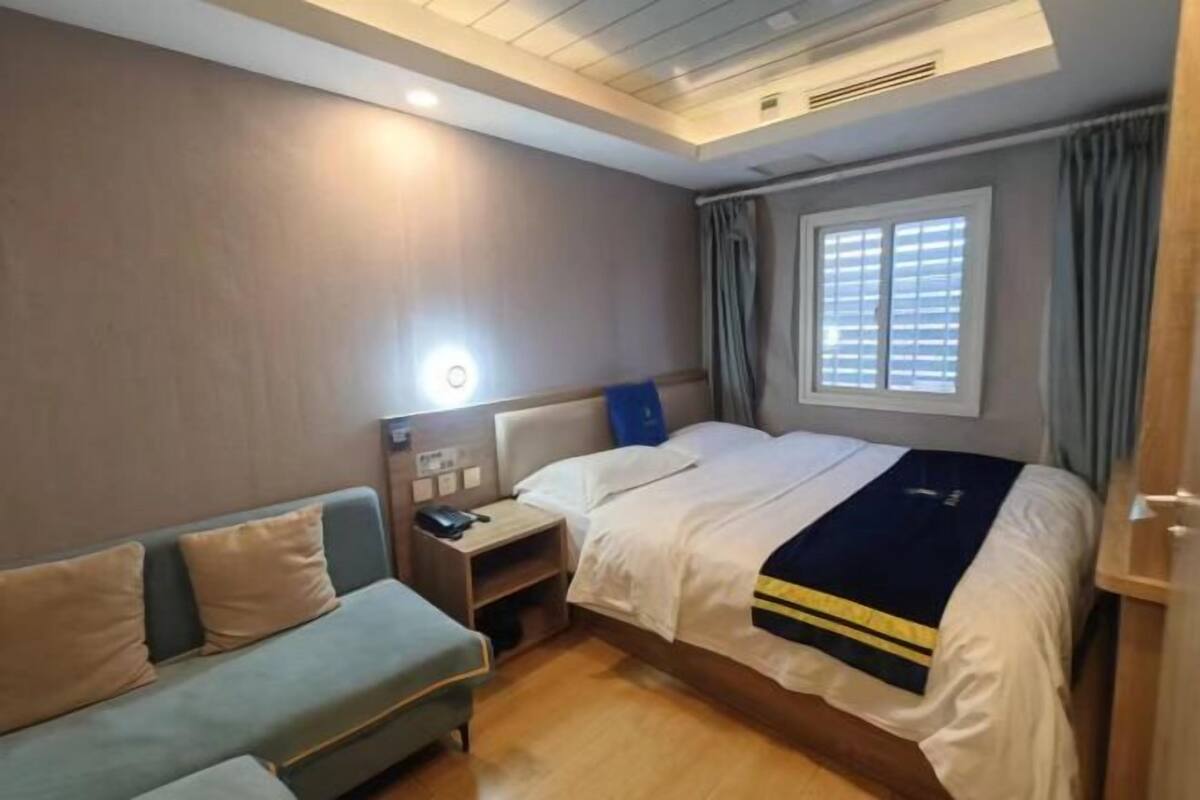 A hotel room with a bed, a sofa, a bedside table with a phone, a window with curtains, and a ceiling light.
