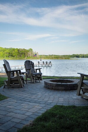 An outdoor seating area with Adirondack chairs and a fire pit, overlooking a calm lake with a dock in the distance.
