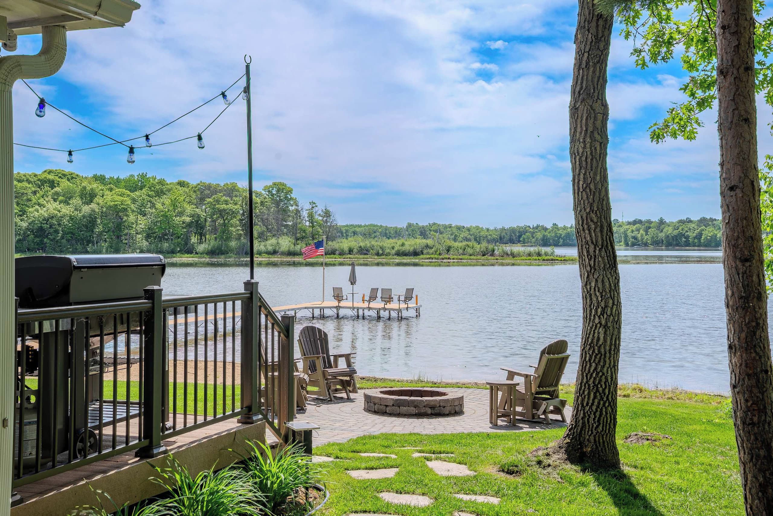 A lakeside deck with outdoor seating and a grill, overlooking a calm lake and forested area.