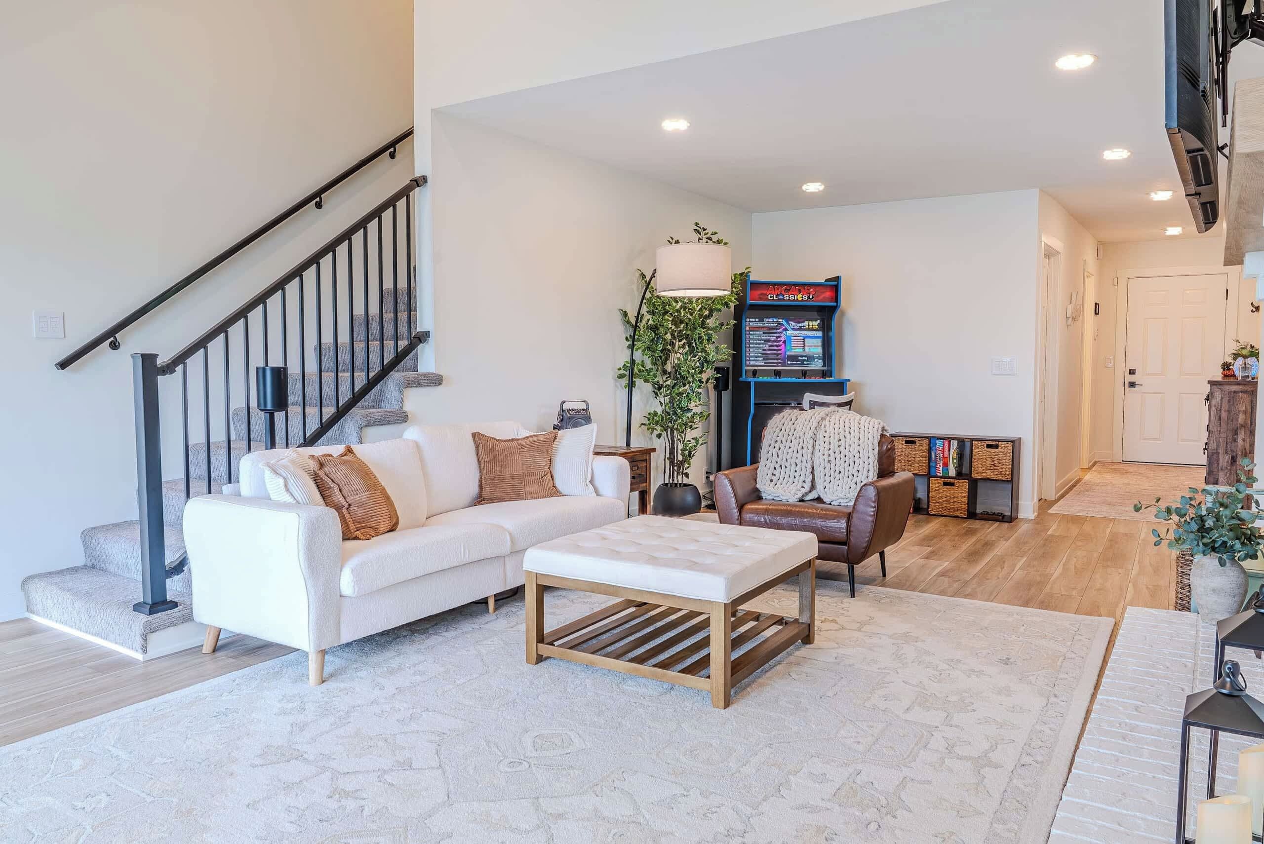 A modern living room with a staircase, a sofa set, a coffee table, a gaming console, and a bookshelf.