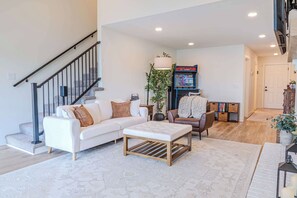 A modern living room with a staircase, a sofa set, a coffee table, a gaming console, and a bookshelf. - Lakefront Cabin w/ Private Beach, Hot Tub & Sauna (Brainerd)