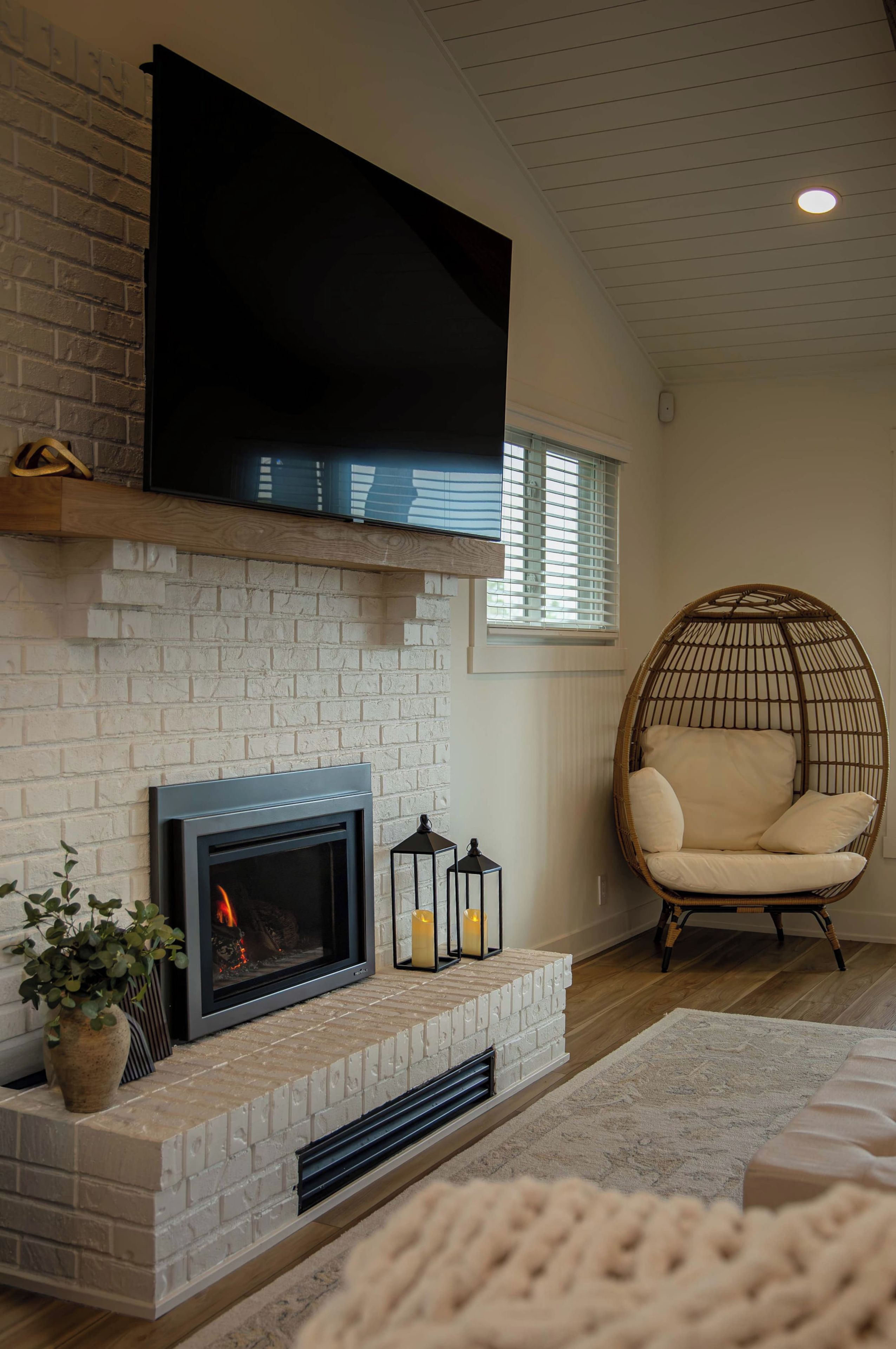 A living room with a fireplace, a television mounted above it, a wicker chair, and a window with blinds.