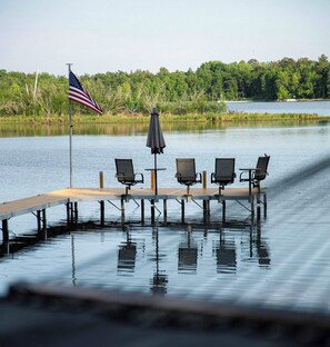A dock with four chairs and an umbrella, a flag, and a calm body of water. - Lakefront Cabin w/ Private Beach, Hot Tub & Sauna (Brainerd)
